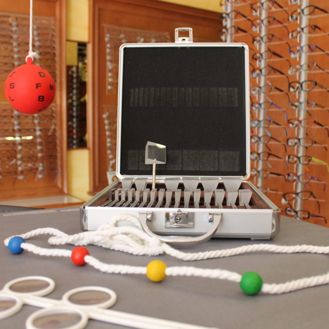 Material para terapia visual Maletín con prismas ópticos, junto a una cuerda con bolas de colores, una bola roja con letras negras y un flipper acomodativo.