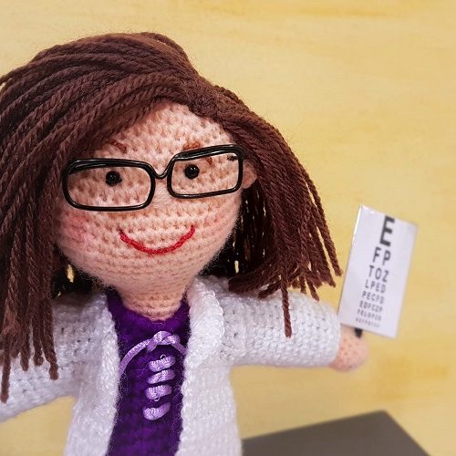 Optometrista Muñeca de lana con cabello castaño, gafas y un optotipo en la mano representando una optometrista.