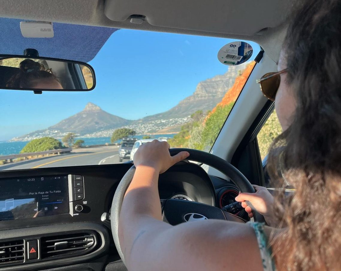 Visión en la conducción Una persona conduciendo un coche con vistas a montañas y mar, utilizando gafas de sol para mejorar su confort conduciendo.